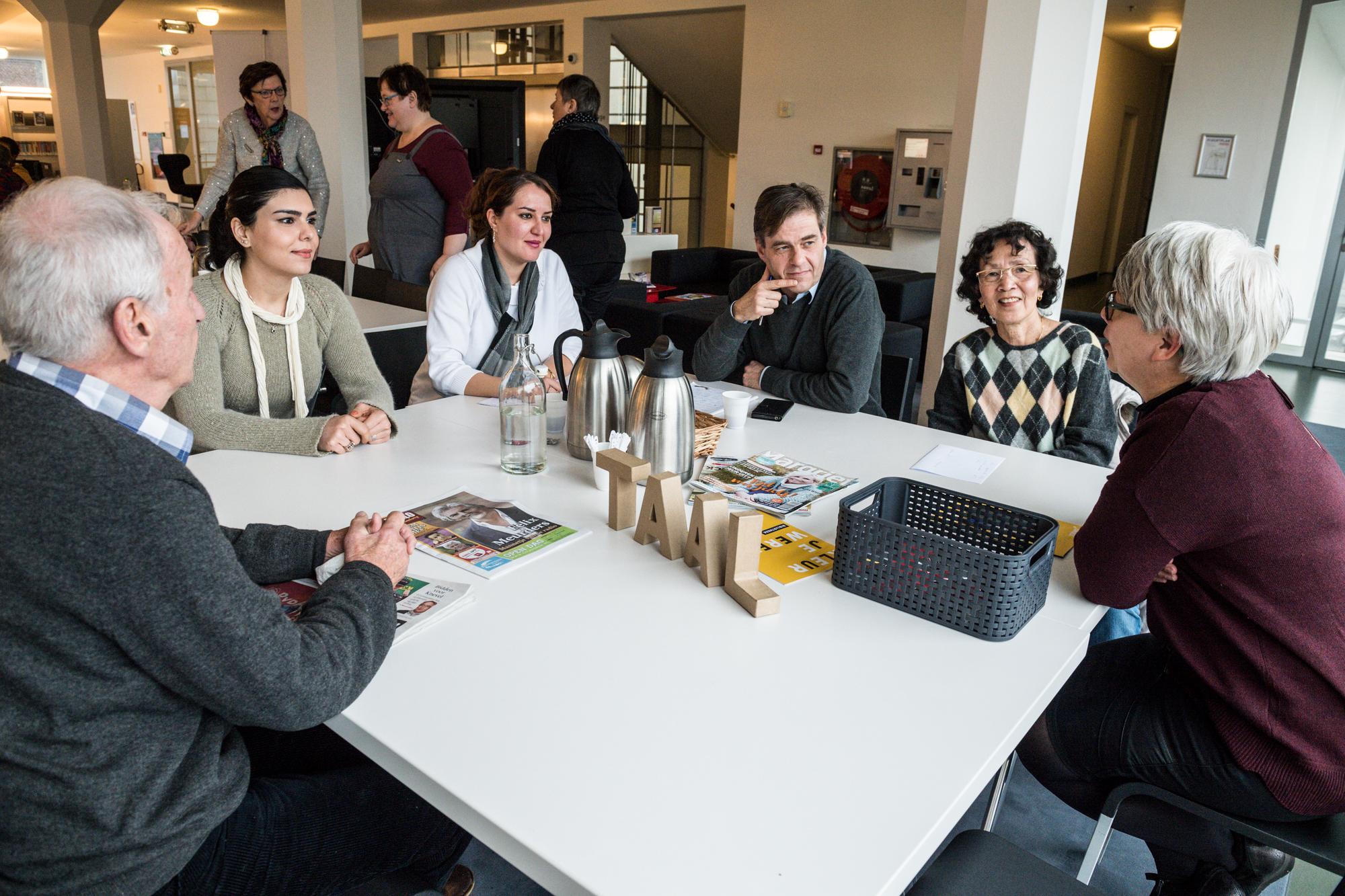 Een groep van zes volwassenen zit samen aan een tafel in gesprek in een lichte ontmoetingsruimte. Op tafel staan koffiekanen, glazen water, tijdschriften en houten letters die samen ‘Taal’ vormen.