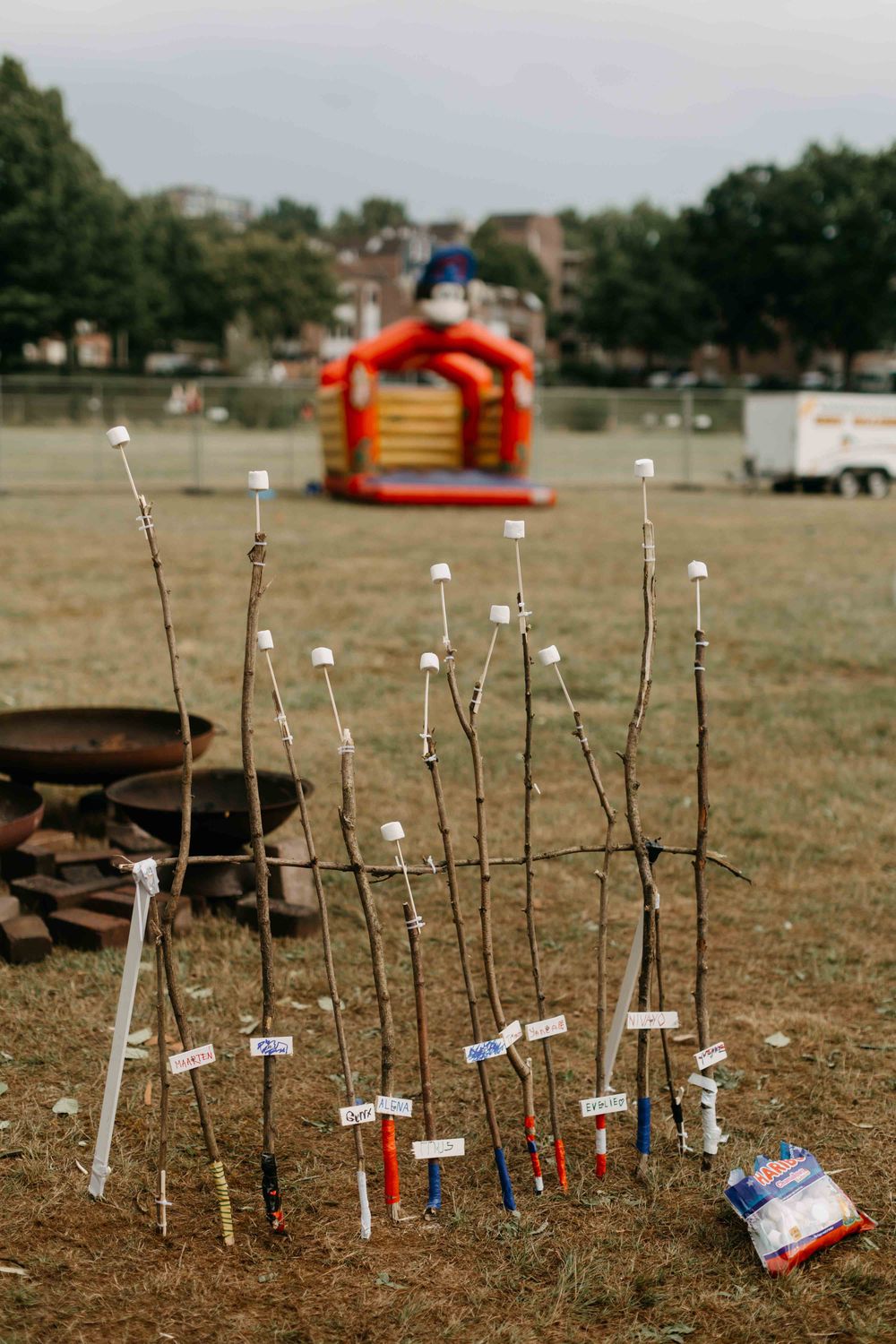  Zelfgemaakte stokken met marshmallows, elk gemarkeerd met een naam, staan opgesteld in het gras, met op de achtergrond een springkussen en een zak marshmallows op de grond.