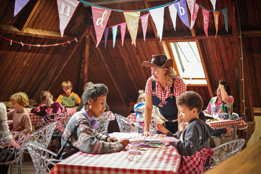 Kinderen zitten aan rood-wit geblokte tafels te tekenen en knutselen tijdens het kinderboekenfeestje van Bob Popcorn in een sfeervolle houten zolderruimte. Een begeleider in cowboyhoed helpt een kind aan tafel terwijl kleurrijke slingers boven de groep hangen.
