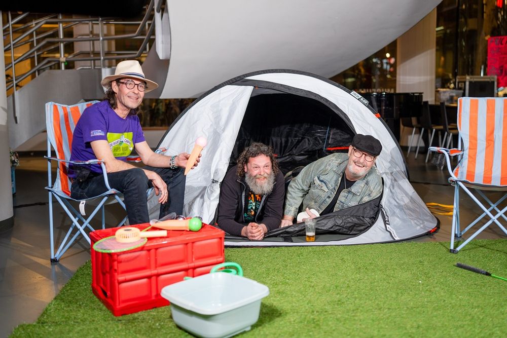 Drie mannen poseren in een speelse kampeeropstelling tijdens Frictions Festival #12, binnen een moderne ruimte; twee liggen lachend in een grijze tent met een drankje, terwijl de derde op een oranje‑witte klapstoel zit met een hoed en een paarse T‑shirt, omringd door kampeerspullen zoals een rode krat, een witte teil en speelgoed op een stukje kunstgras.