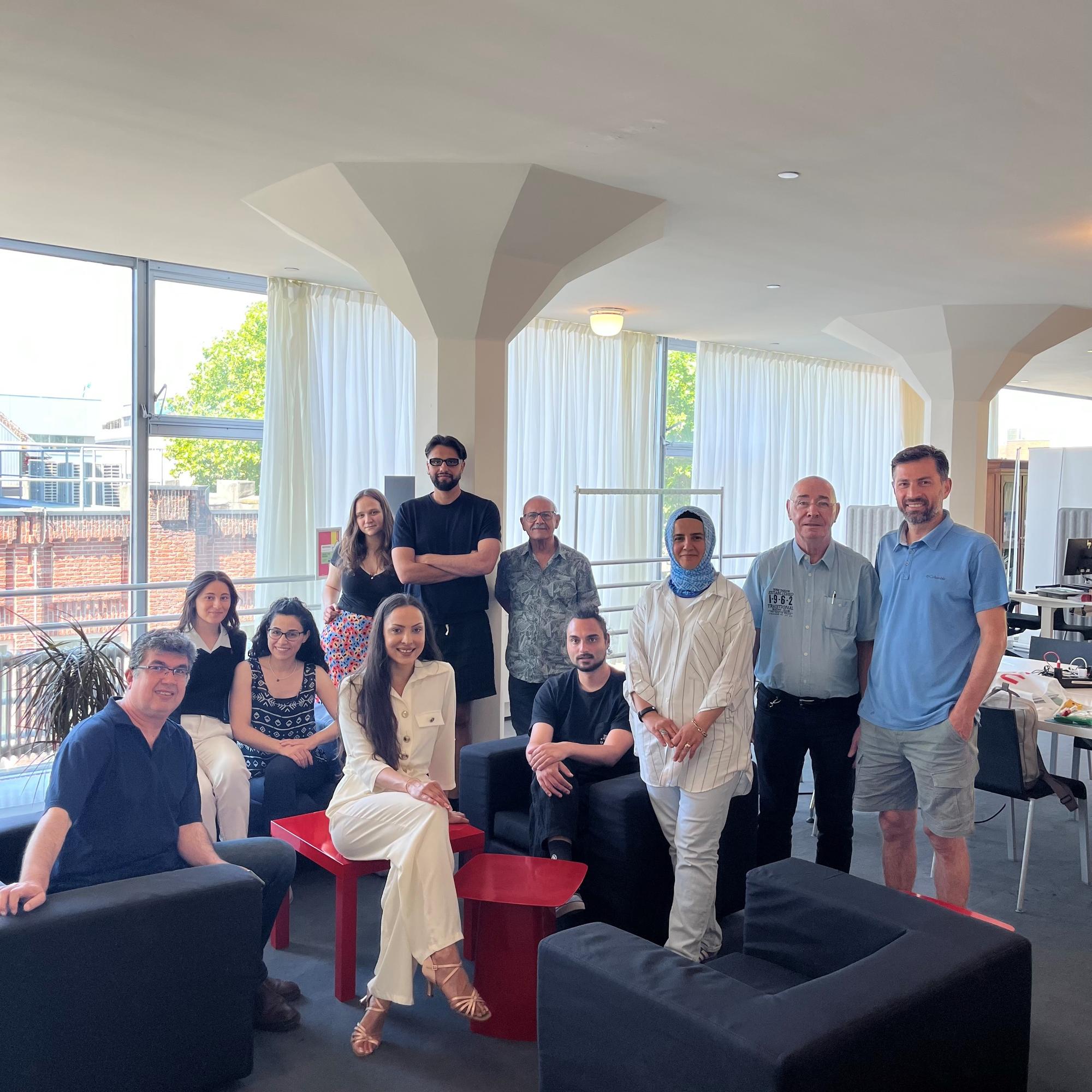 Een groep van twaalf mensen poseert samen in een lichte ruimte met grote ramen en witte gordijnen, zittend en staand rond zwarte stoelen en rode tafeltjes.