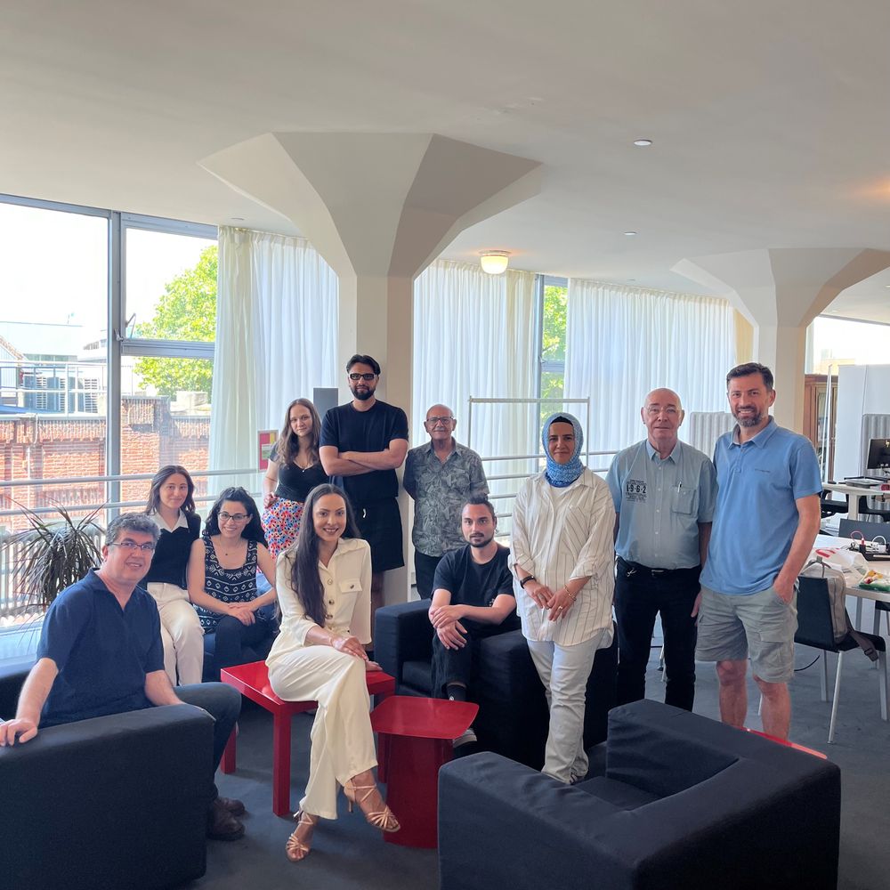 Een groep van twaalf mensen poseert samen in een lichte ruimte met grote ramen en witte gordijnen, zittend en staand rond zwarte stoelen en rode tafeltjes.