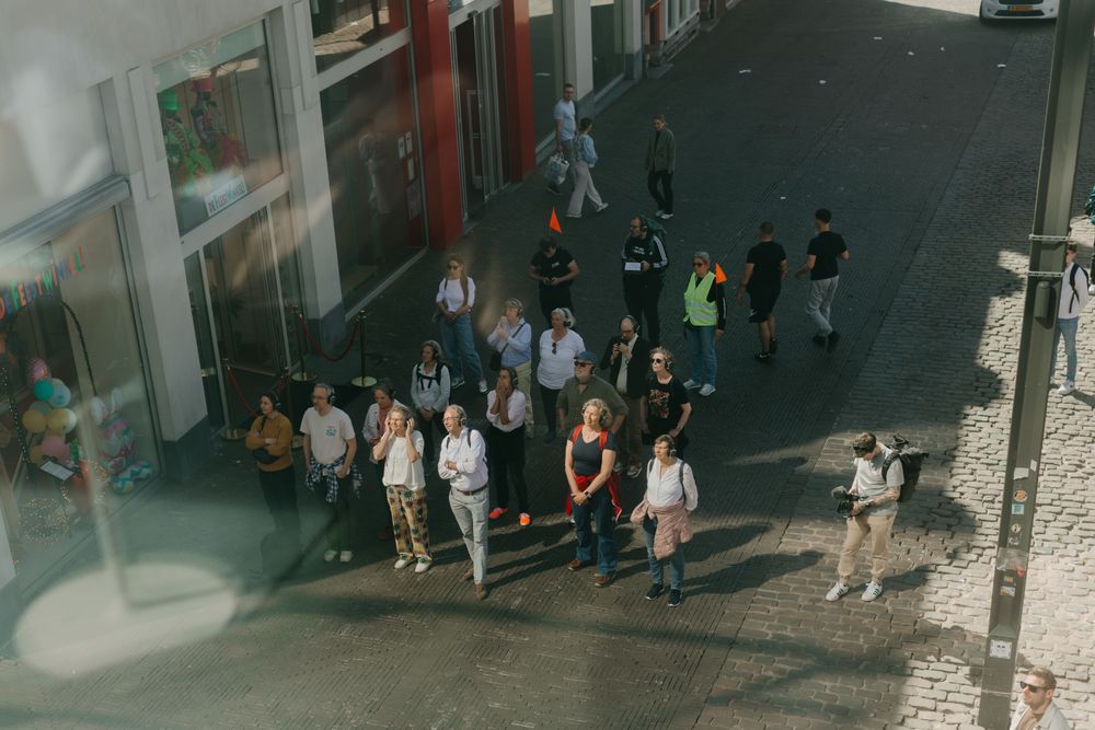 Groep mensen die op een straat samenkomen en omhoog kijken richting SCHUNCK Glaspaleis, gezien van bovenaf door een raam.
