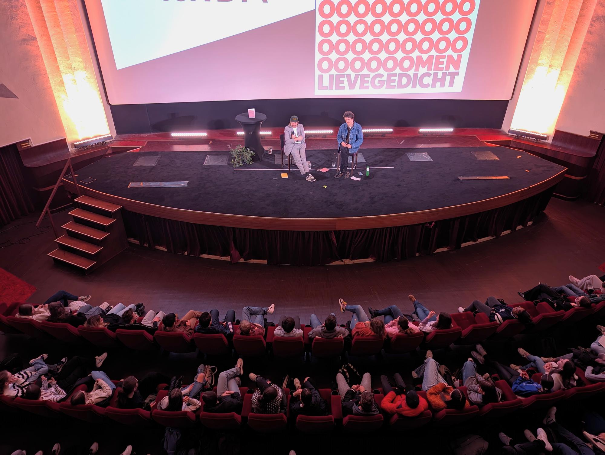 Schunck Poeziefestival 2024 Filmzaal in Royal Theater Heerlen gevuld met jongeren die luisteren naar twee sprekers op een groot podium, met op de achtergrond een scherm waarop “Lievegedicht” en “Oooooooooomen” te zien is in rode en witte letters, in een sfeervol verlichte omgeving.