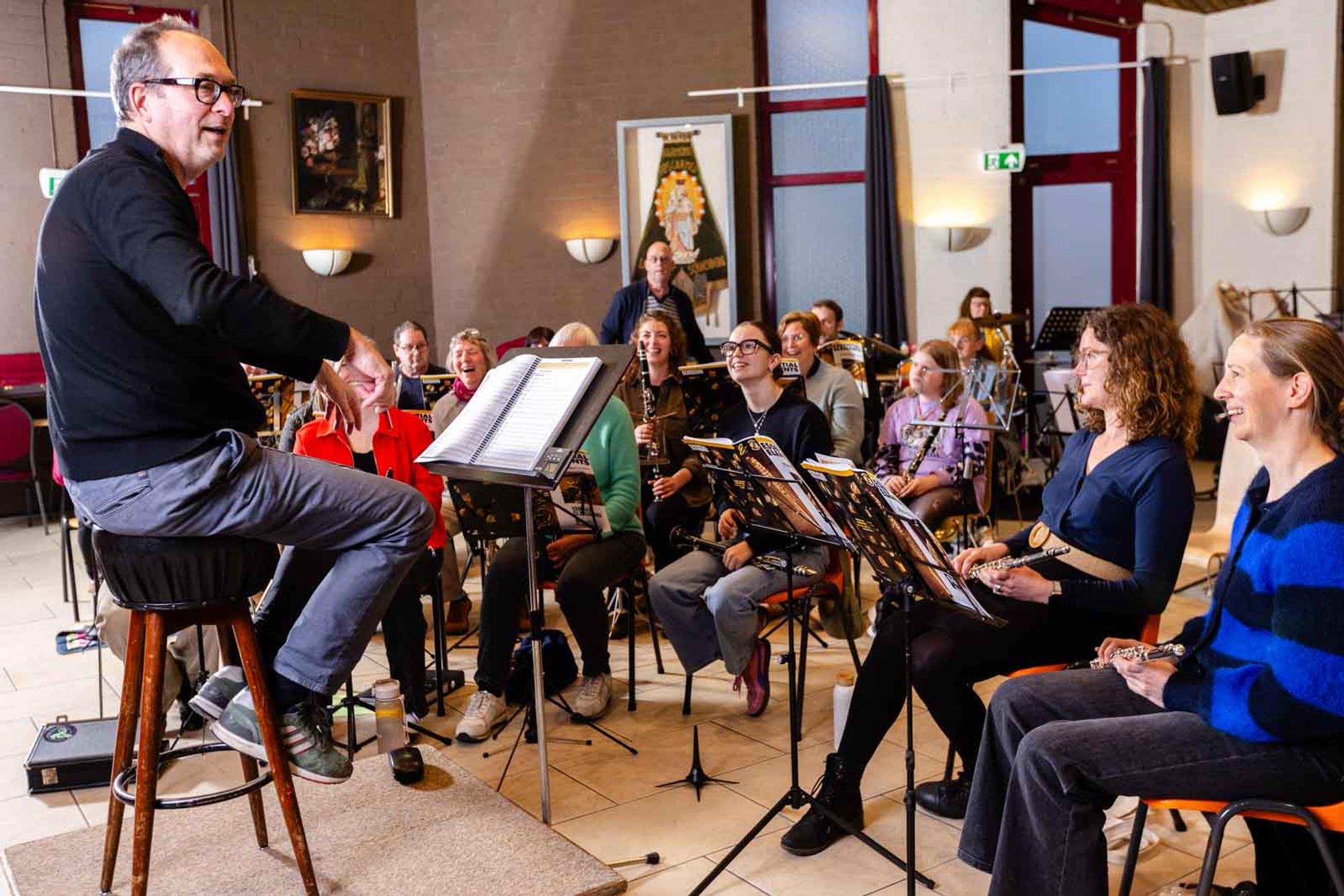 Dirigent leidt een groep volwassen muzikanten tijdens een repetitie in een lichte zaal. De deelnemers zitten met blaasinstrumenten achter muziekstandaards en luisteren aandachtig terwijl er zichtbaar een ontspannen en enthousiaste sfeer heerst.