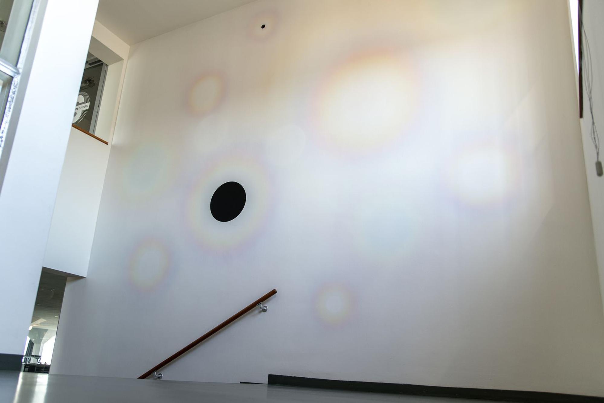 Minimalistische muurschildering in SCHUNCK met subtiele pastelkleurige cirkels verspreid over een witte muur en een grote zwarte stip in het midden, zichtbaar vanuit het trappenhuis.