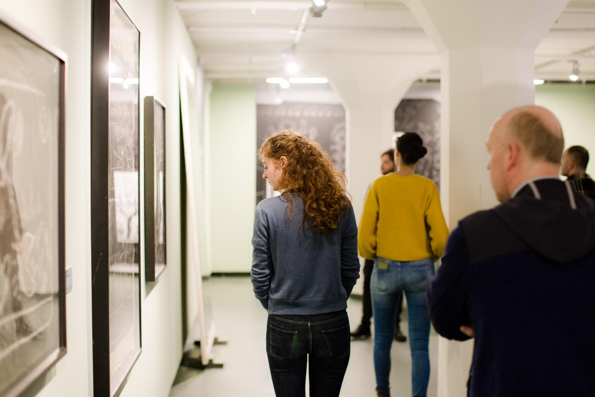Een groep mensen loopt door een kunstgalerij of tentoonstelling. Er hangen ingelijste kunstwerken aan de muren en de mensen bekijken deze. De ruimte is binnen met witte muren en plafondverlichting.