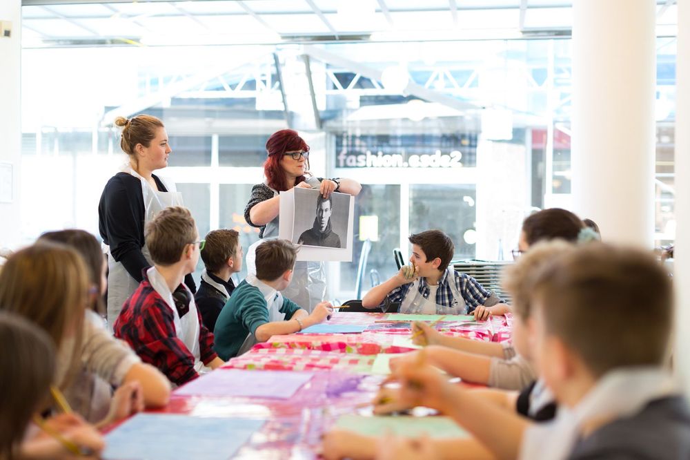 Kinderen zitten aan een lange tafel met verfmatten terwijl een vrouw voor de klas een zwart-wit portret omhooghoudt; naast haar staat een andere begeleider en de kinderen kijken aandachtig naar het portret.
