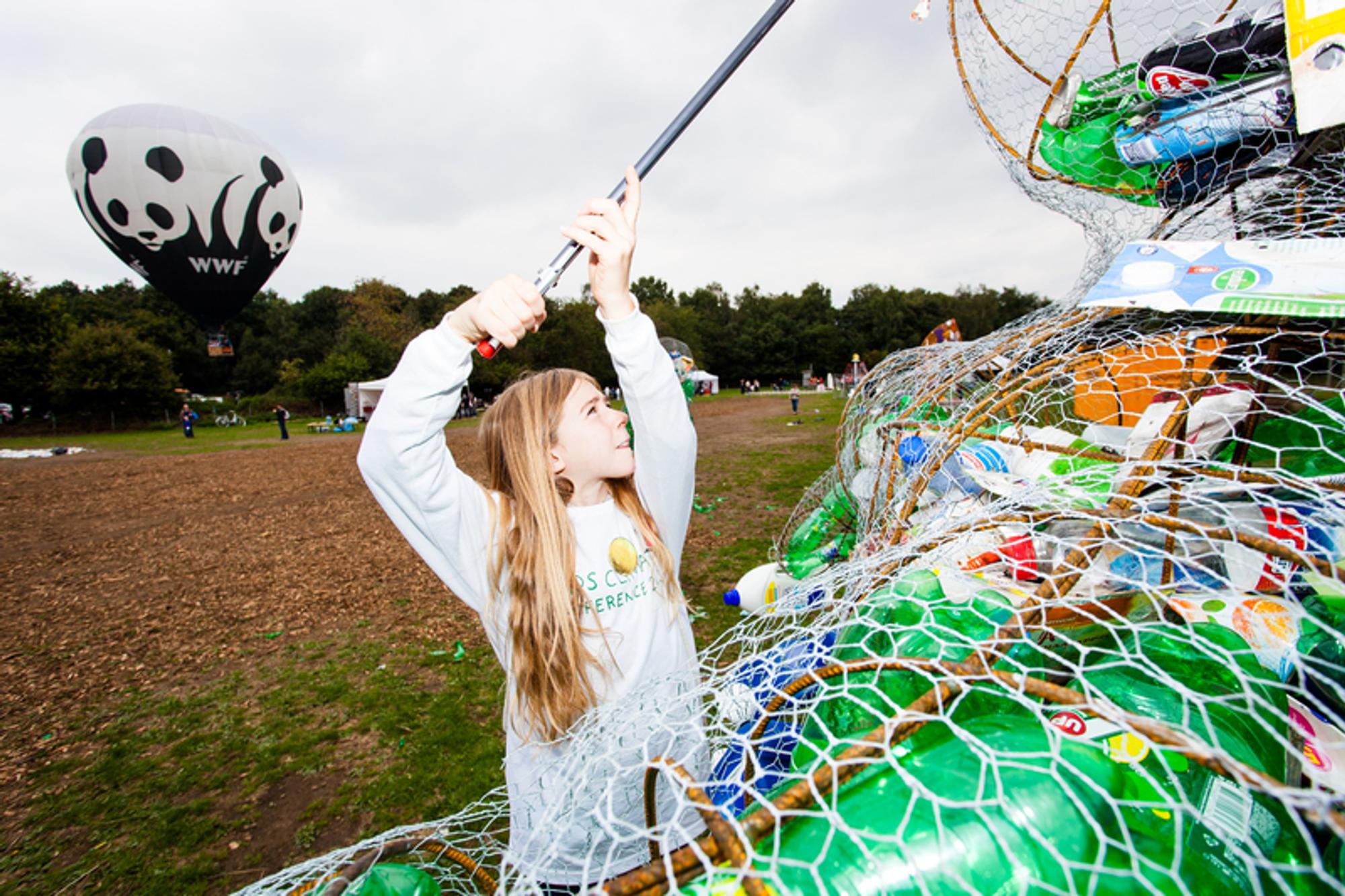 Een meisje dat staat bij een aantal netten met plastic-afval erin, terwijl ze met een stok iets vasthoud dat zich buiten het beeld bevindt. Op de achtergrond is een luchtballon van WWF te zien. 