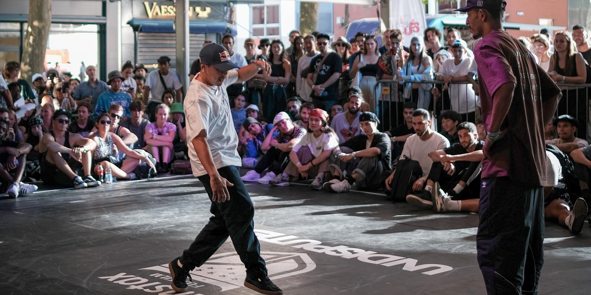 Twee dansers staan tegenover elkaar in een spannende breakdance battle, omringd door een grote, aandachtige menigte die in een cirkel zit en staat rondom de dansvloer.