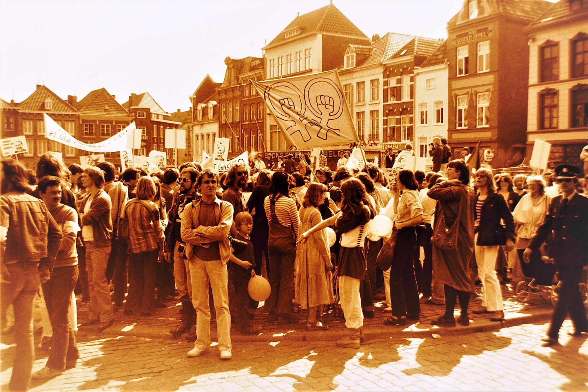 Demonstratie op een stadsplein met een grote menigte en spandoeken, waaronder een vrouwensymbool, tegen een achtergrond van historische gebouwen.