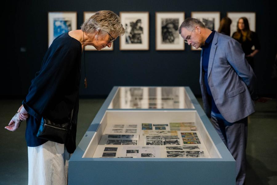 Twee bezoekers bestuderen documenten en archiefmateriaal in een vitrinetafel tijdens een tentoonstelling, met ingelijste foto's aan de wand op de achtergrond.