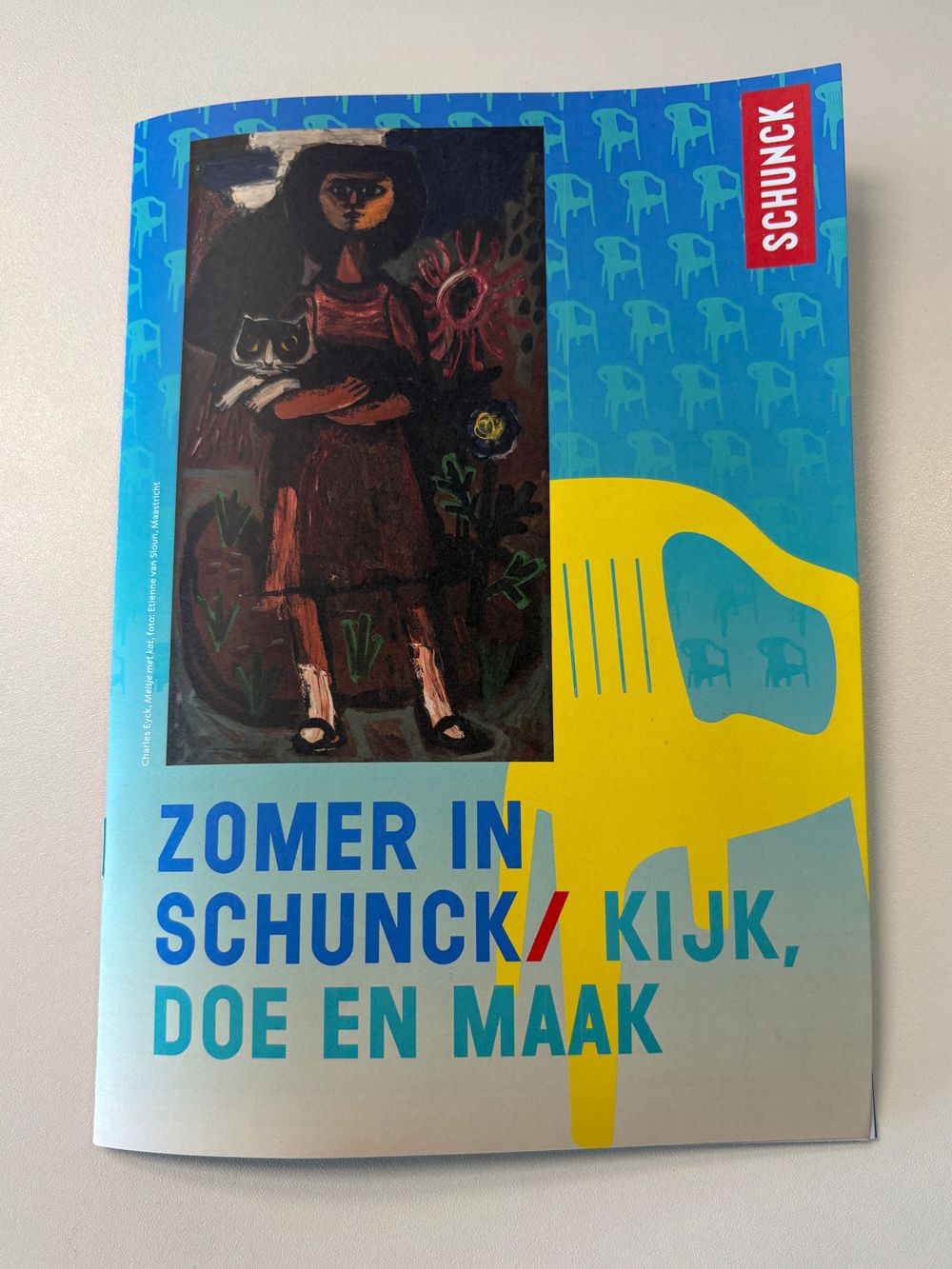 Flyer met blauwe en gele achtergrond en een illustratie van een persoon die een kat vasthoudt, omringd door abstracte elementen. De tekst luidt: 'ZOMER IN SCHUNCK / KIJK, DOE EN MAAK'. Rechts staat 'SCHUNCK' verticaal op een rode strook.
