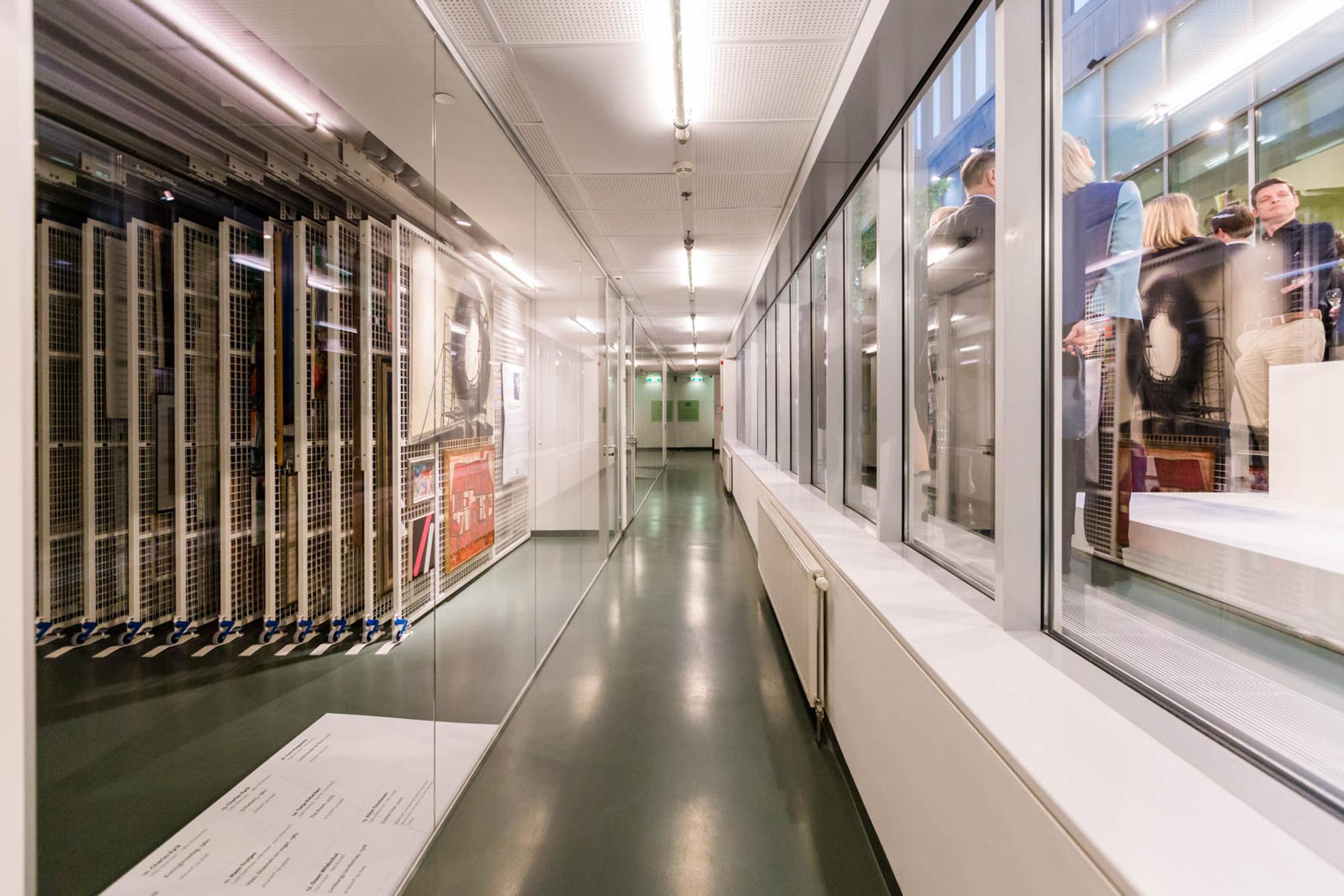 De gang van het kunstdepot, met links achter glas de rekken waar schilderijen hangen en rechts het atrium staat een groep mensen.