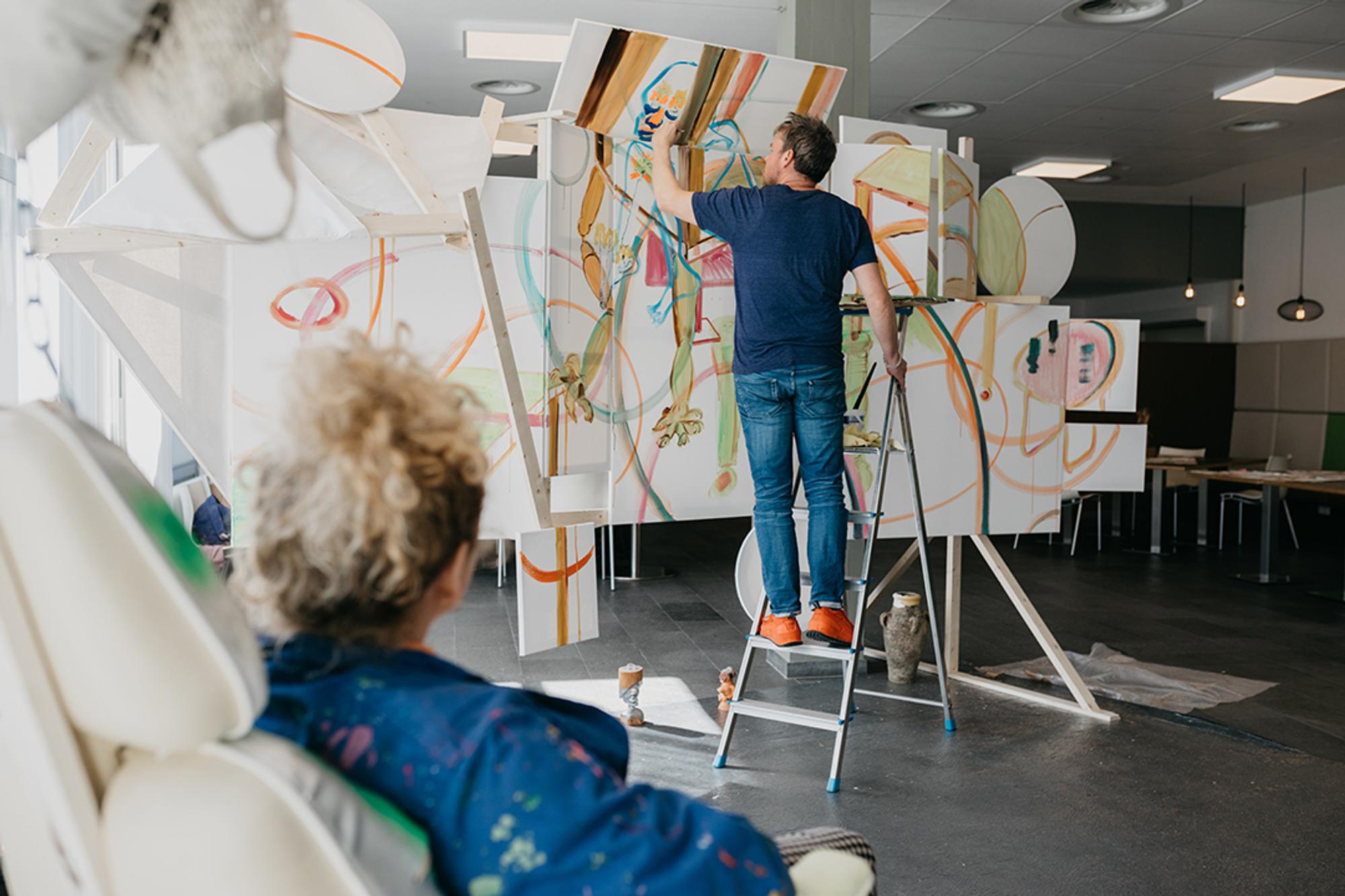 David Bade in blauw shirt staat op een ladder en schildert op een doek van zijn kunstinstallatie. Op de voorgrond zit een vrouw in een tandartsstoel.