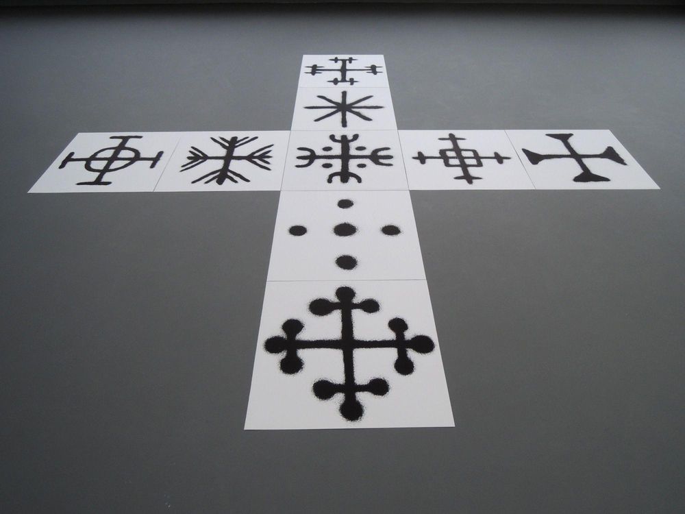 Op witte panelen liggen zwarte symbolen en tekens, in de vorm van een groot kruis op de vloer gerangschikt. De vormen lijken ritueel of grafisch en variëren van kruizen tot sterachtige patronen.