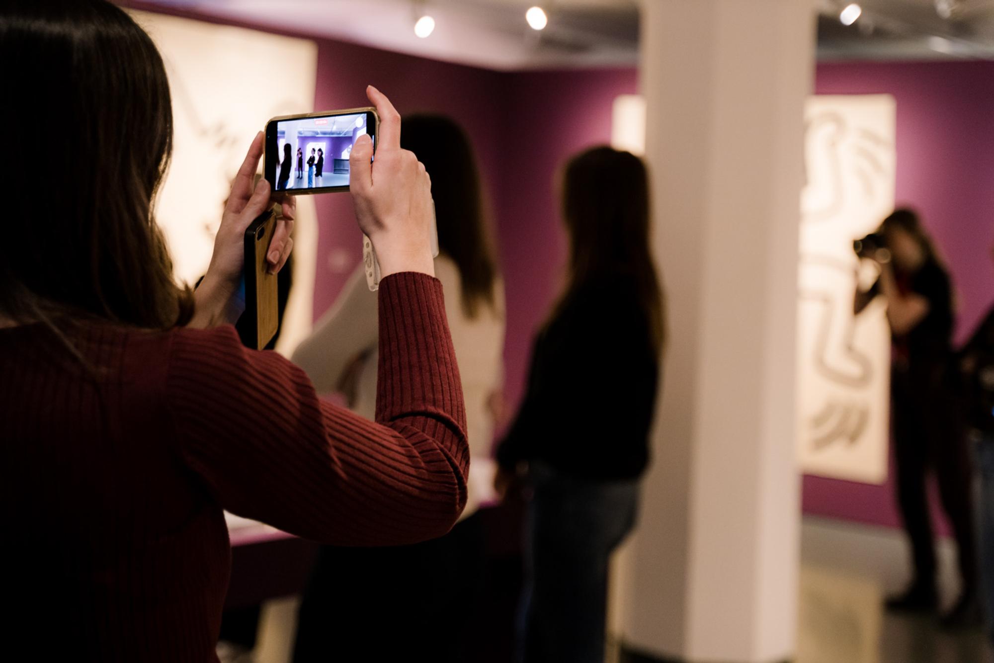 Een groep mensen in een kunstgalerie. Eén persoon op de voorgrond maakt een foto met een smartphone, terwijl anderen de kunstwerken aan de muren bekijken. De achtergrond heeft grote, abstracte schilderijen of tekeningen op paarse muren.