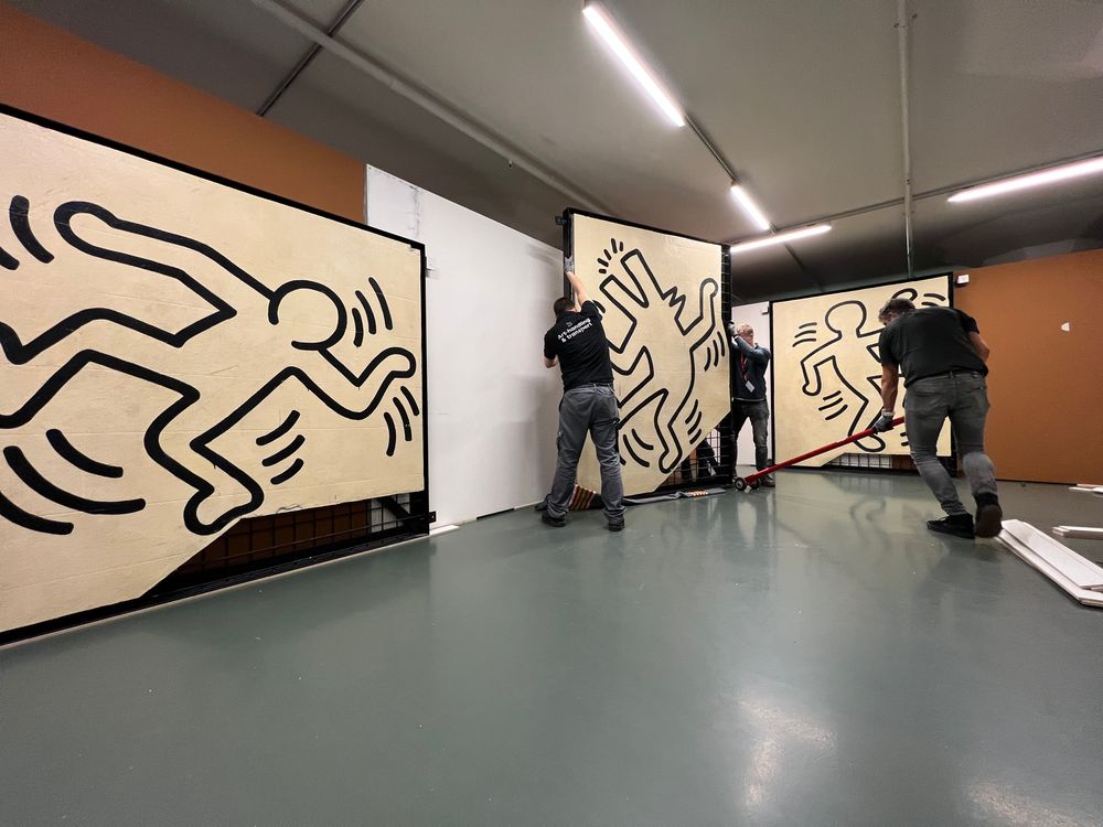 Drie mannen installeren grote muurdelen met iconische zwarte figuren van Keith Haring in een tentoonstellingsruimte bij SCHUNCK.
