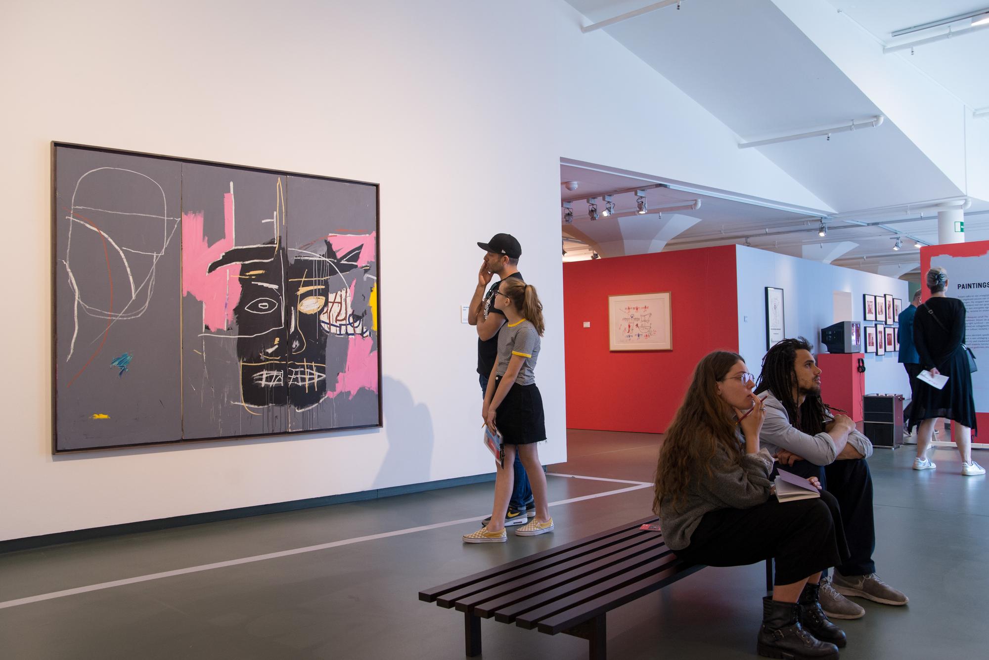 Bezoekers bekijken aandachtig een kunstwerk van Jean-Michel Basquiat in een museumzaal met witte muren en rode accenten.