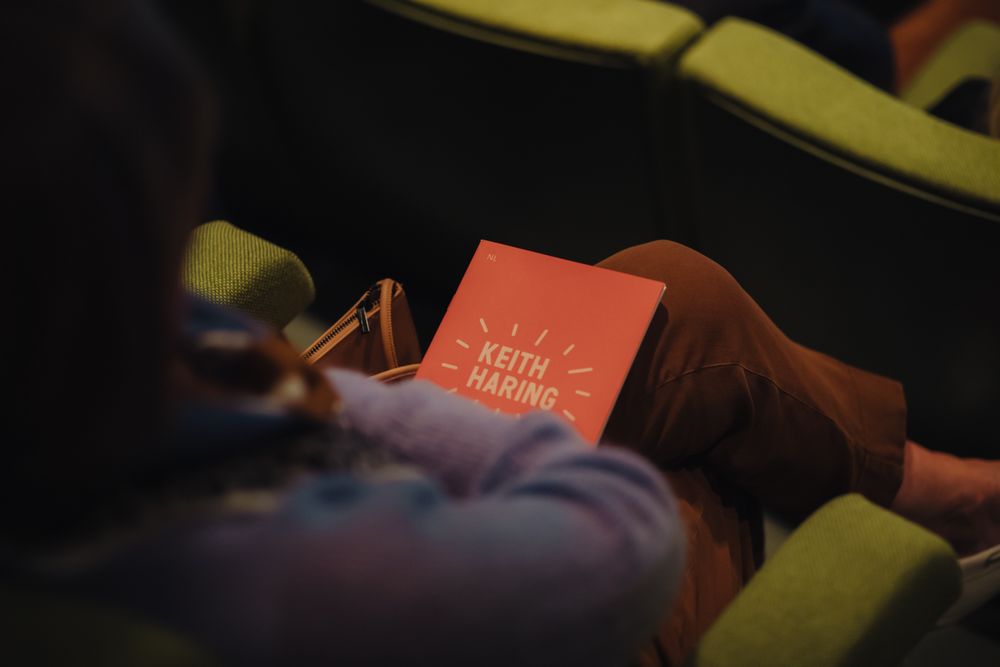 Persoon zit in een groene theaterstoel en houdt een rood boekje vast met de titel "Keith Haring" in witte letters en stralende lijnen eromheen.
