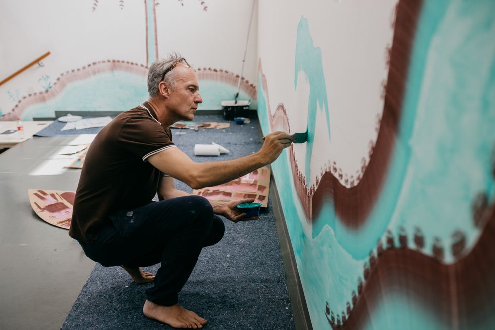 Kunstenaar Gijs Frieling schildert op blote voeten een muurschildering op de muur van SCHUNCK in Heerlen, zittend op zijn hurken en werkend aan een golvend patroon in groen en bruin.