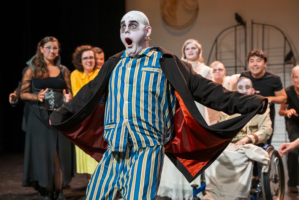 Groep mensen op een toneel tijdens een theatervoorstelling. Centraal staat iemand in gestreepte pyjama’s met een open zwarte jas met rode voering. Op de achtergrond staan en zitten anderen in diverse kostuums, waaronder formele en alledaagse kleding. Eén persoon zit in een rolstoel. Gezichten zijn onherkenbaar gemaakt.