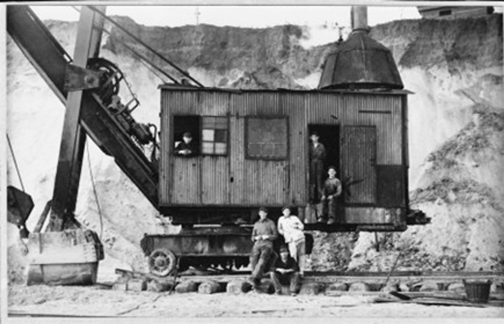 Oude industriële graafmachine met grote bak in een steengroeve, met vijf mannen: twee bij de cabine en drie op de grond, tegen een achtergrond van een steile rotswand.
