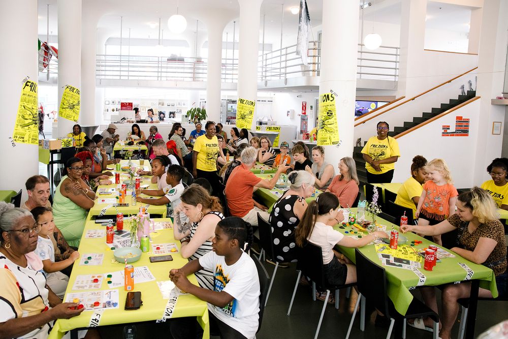 Een grote groep mensen van verschillende leeftijden zit aan lange tafels met groene tafelkleden en speelt het spel Bon Ku Ne tijdens Frictions Festival #9; vrijwilligers in gele T‑shirts lopen rond, terwijl deelnemers lachen, praten en geconcentreerd hun kaarten invullen in een lichte zaal met witte zuilen en gele posters aan de muren.