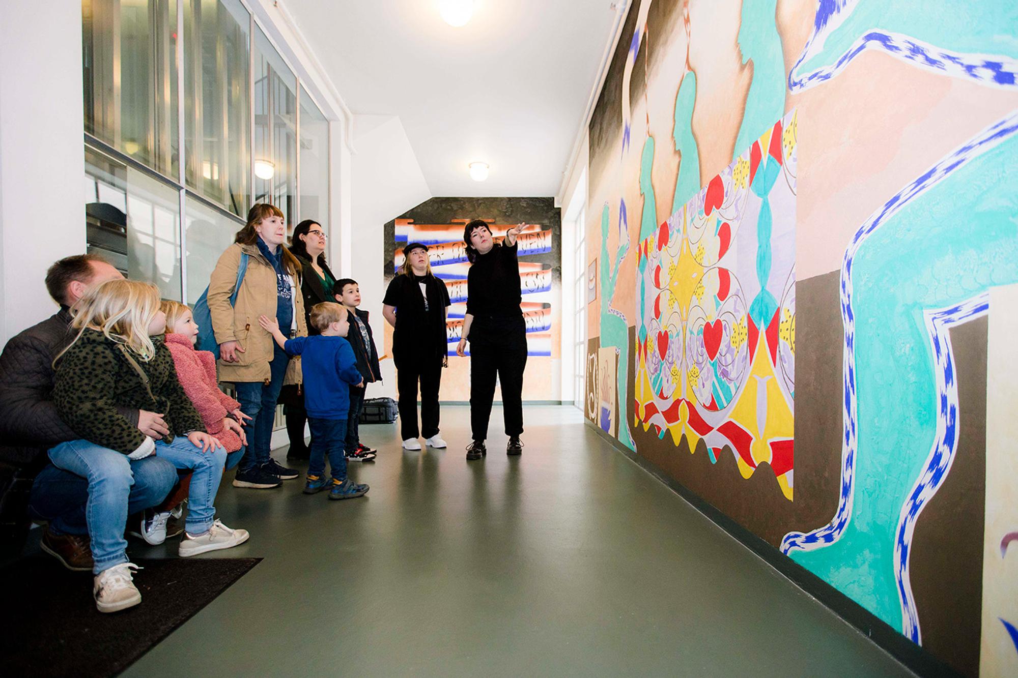 Groep volwassenen en kinderen in een gang of corridor, staand en zittend terwijl ze een groot, kleurrijk en abstract muurkunstwerk bekijken. De ruimte is goed verlicht met plafondlampen en heeft ramen aan één zijde.