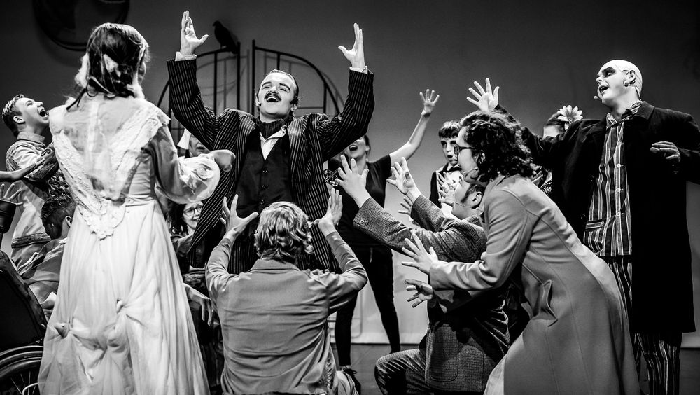 Zwart-witfoto van een groep mensen in theatrale kostuums tijdens een expressieve toneelvoorstelling. Centraal staat iemand in een gestreept pak met opgeheven armen, omringd door anderen die reageren met uitgesproken gebaren. Gezichten zijn onherkenbaar gemaakt.