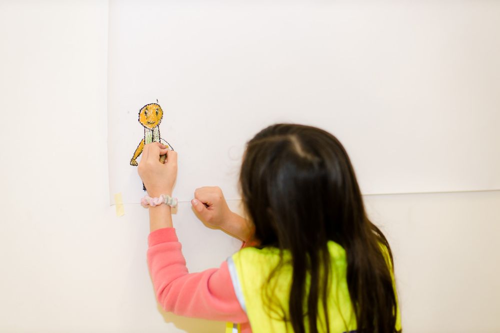 Meisje in een geel hesje tekent een poppetje op de muur.