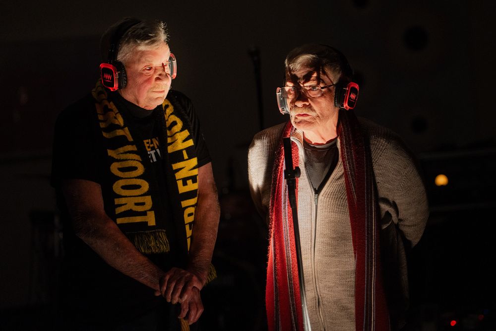 Twee oudere mannen staan in warm licht bij een microfoon, beiden dragen rode koptelefoons; links Sven Vek met een zwarte shirt en gele sjaal, rechts Jeffrey Hanssen met bril, grijs vest en rode sjaal, tijdens een optreden van IMP SYN waarin poëzie en elektronica samenkomen tot een rauwe, experimentele performance.