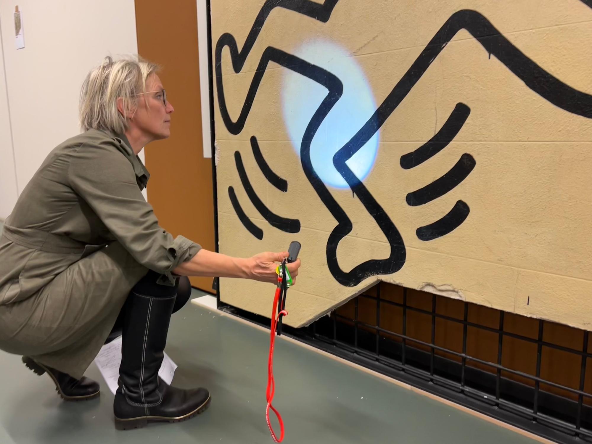Een vrouw inspecteert een muurschildering van Keith Haring met een zaklamp, waarbij ze aandachtig naar details in de zwarte lijnen kijkt.