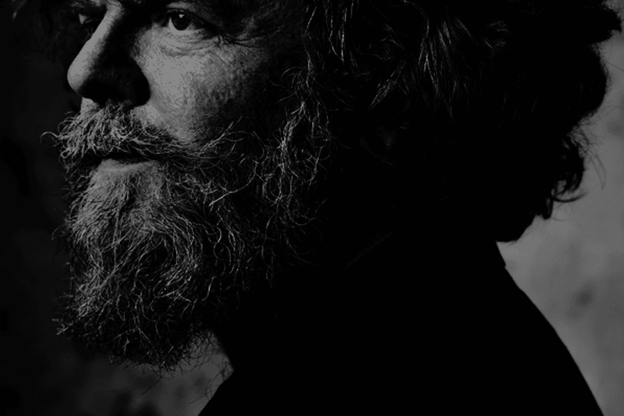 Zwart-wit portret van een man met halflang haar en een volle baard, in profiel gefotografeerd. Het licht valt van opzij en benadrukt de structuur van zijn gezicht tegen een donkere achtergrond.