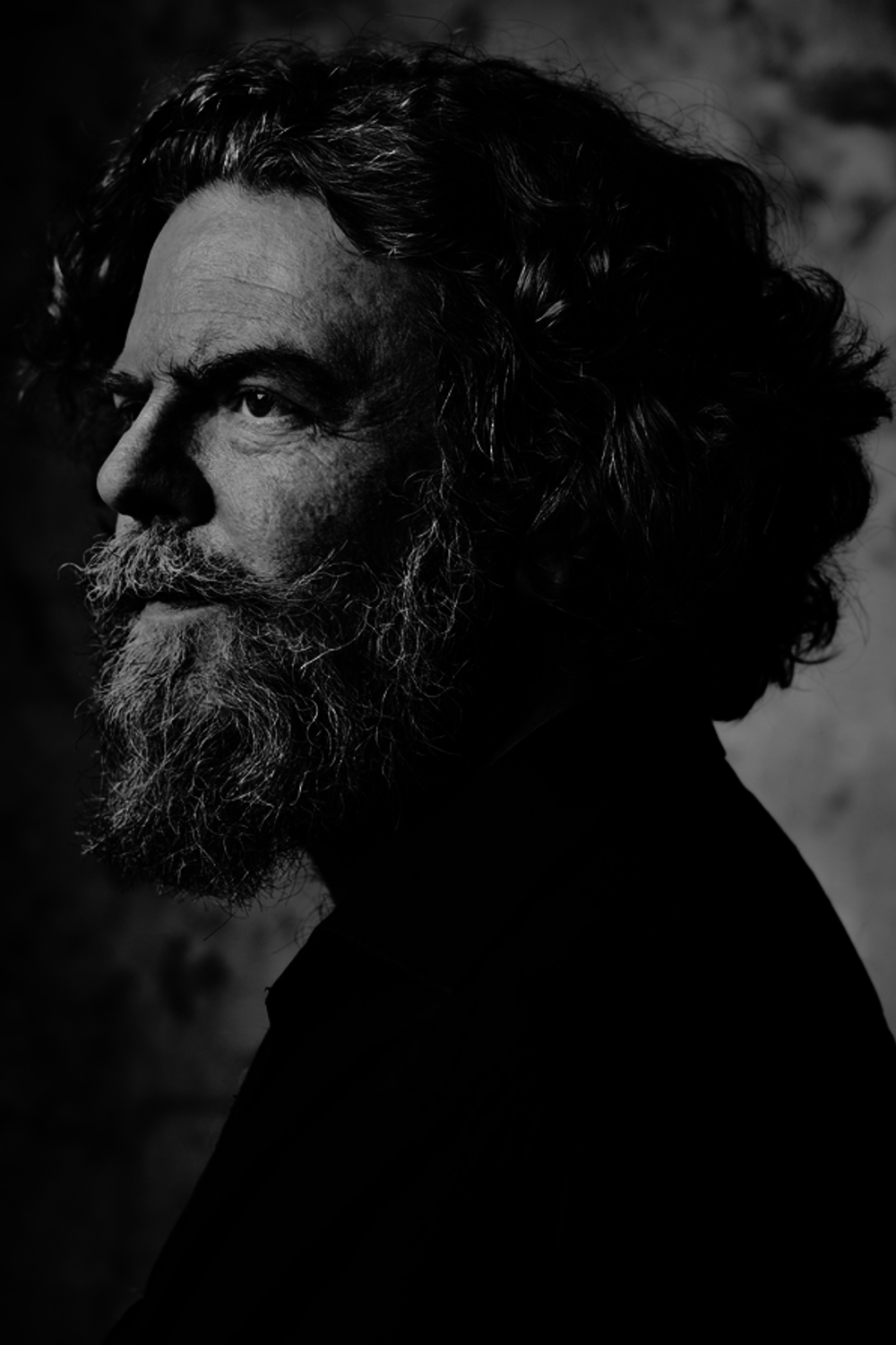 Zwart-wit portret van een man met halflang haar en een volle baard, in profiel gefotografeerd. Het licht valt van opzij en benadrukt de structuur van zijn gezicht tegen een donkere achtergrond.