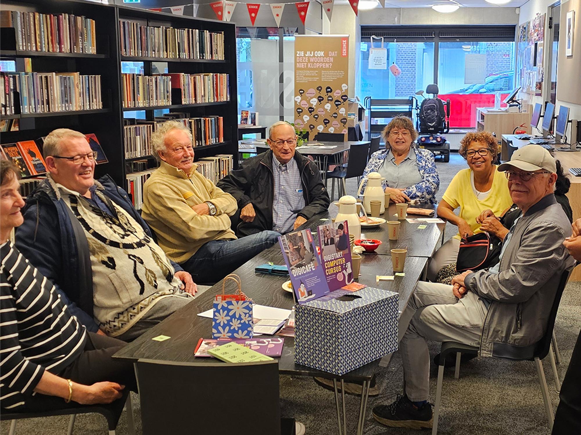 Mensen zitten rond een tafel in de bibliotheek. Op de achtergrond zijn boeken te zien. De mensen lachen