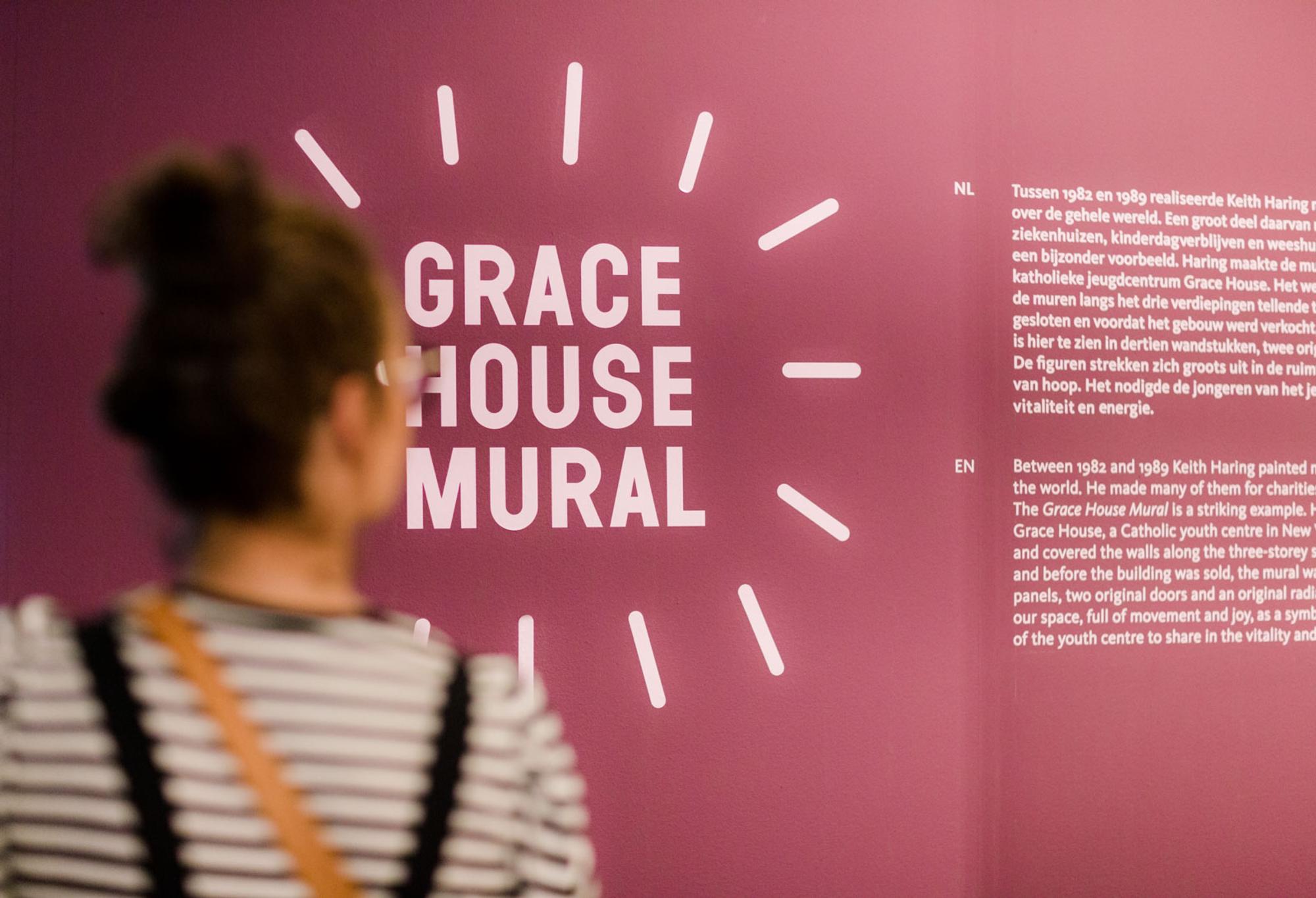 Bezoeker kijkt naar een paarse muur met informatie over de Grace House Mural van Keith Haring, met tekst in het Nederlands en Engels.