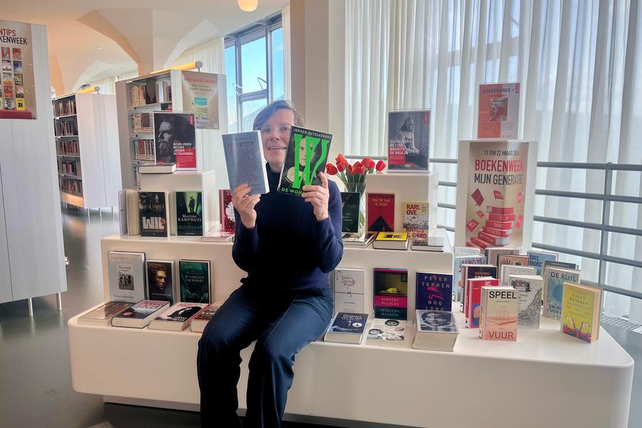 Janna zit met twee boeken in haar handen op een meubel in de bibliotheek. Om haar heen legen meerdere boeken.