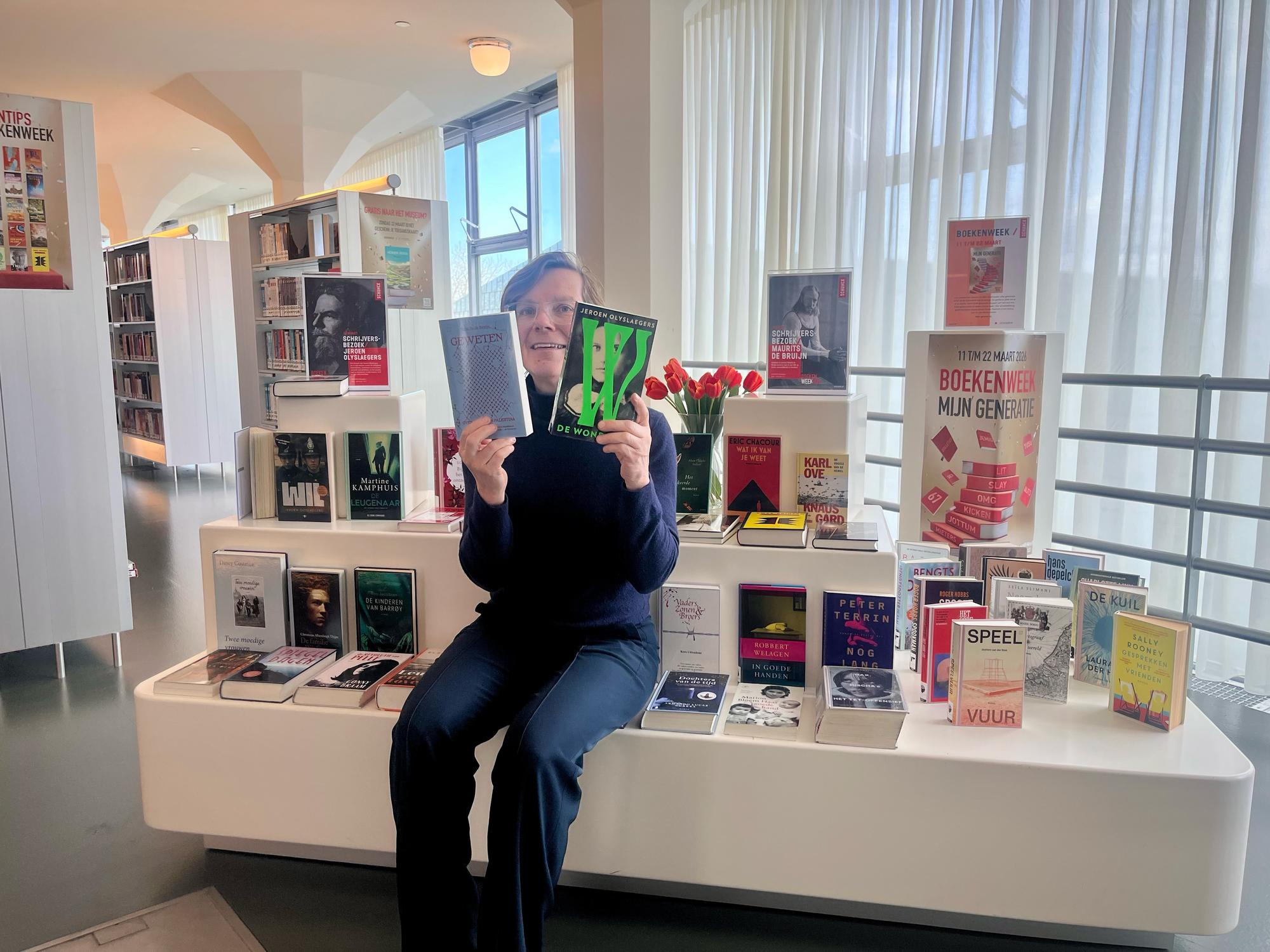 Janna zit met twee boeken in haar handen op een meubel in de bibliotheek. Om haar heen legen meerdere boeken.