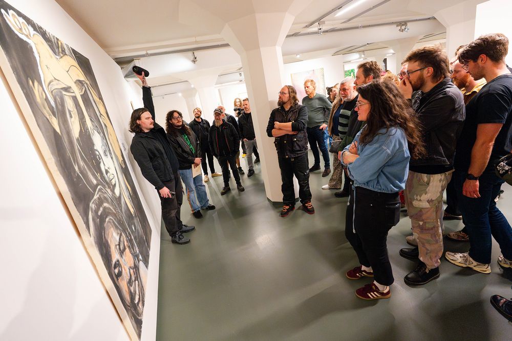 Een groep bezoekers luistert aandachtig naar een man die bij een groot schilderij staat en met één arm omhoog iets uitlegt; de mensen staan in een halve cirkel in een lichte galerie met witte muren en grijze vloer, terwijl het kunstwerk prominent aan de wand hangt. Tijdens Frictions Festival #11 bij SCHUNCK. 