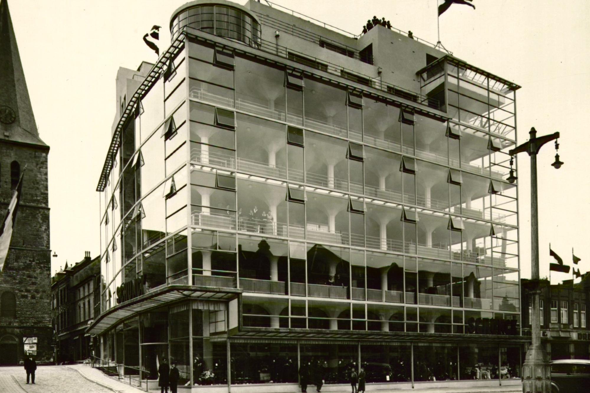 Historische zwart-witfoto van de opening van het Glaspaleis in Heerlen in 1935. Het moderne gebouw met glazen gevel staat op een hoek van een straat met kinderkopjes. Vlaggen wapperen op het dak en mensen zijn zichtbaar rondom het gebouw. Links is een kerkachtig bouwwerk te zien. Onderaan staat in cursief 'Montelplein' en 'L.C.J.S.'.