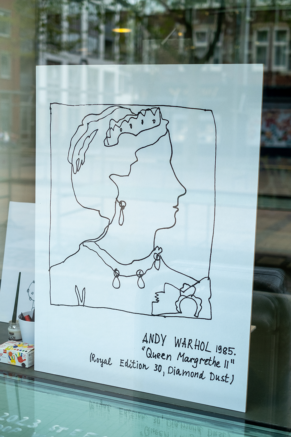 Lijntekening van een vrouw met kroon, oorbel en ketting, getiteld 'Queen Margrethe II' door Andy Warhol (1985), tentoongesteld achter glas.