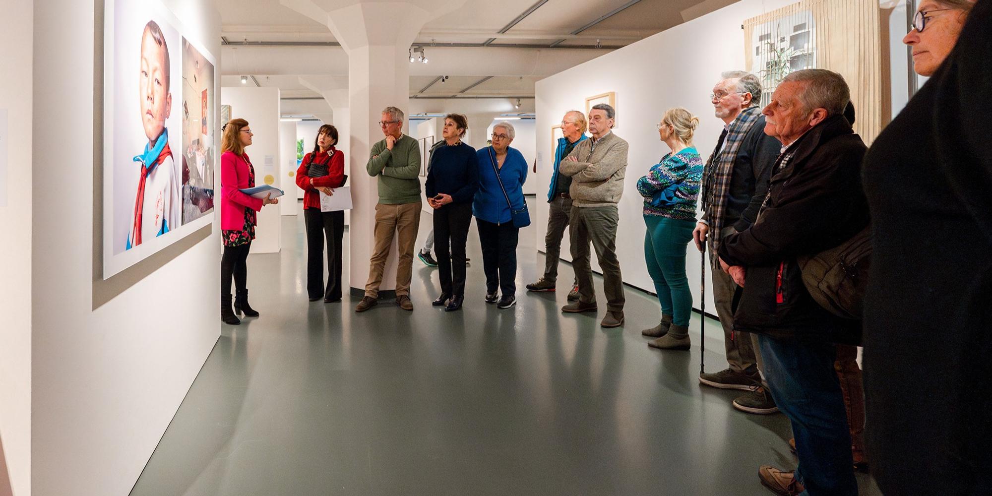 Groep mensen luistert naar een rondleiding in een kunstgalerie. Aan de muur hangen grote portretfoto’s, en een persoon geeft uitleg. De ruimte heeft witte muren, een grijze vloer en moderne verlichting.