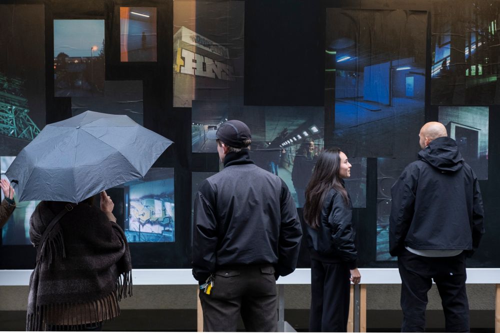 Vier mensen bekijken een groot fotowerk met nachtelijke stedelijke scènes in blauwe en groene tinten. Eén persoon houdt een paraplu vast, terwijl de anderen in gesprek lijken over het kunstwerk.