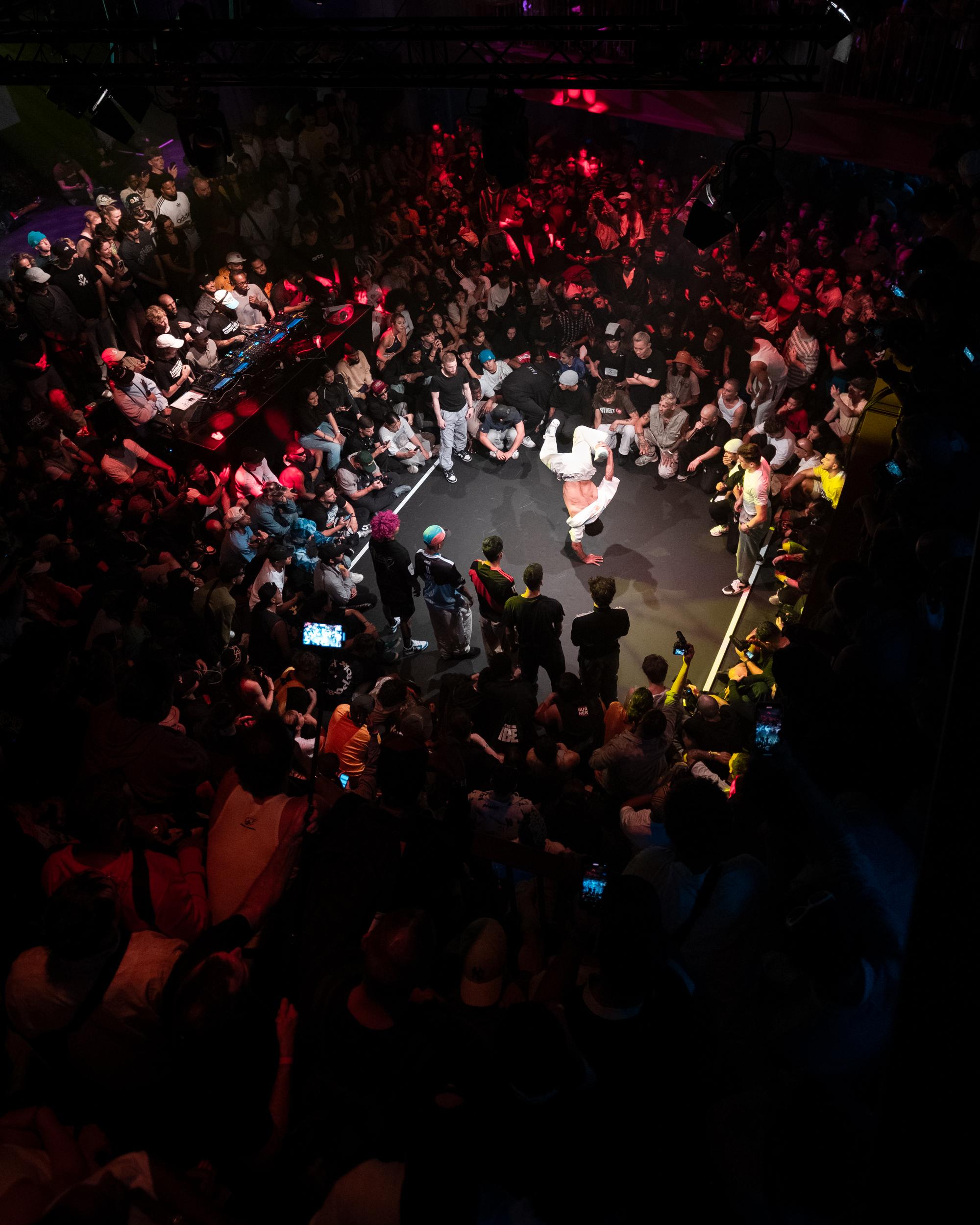 Grote groep mensen staat en zit in een kring in een donkere zaal met felgekleurde verlichting, terwijl in het midden een danser op zijn handen een breakdance move uitvoert.
