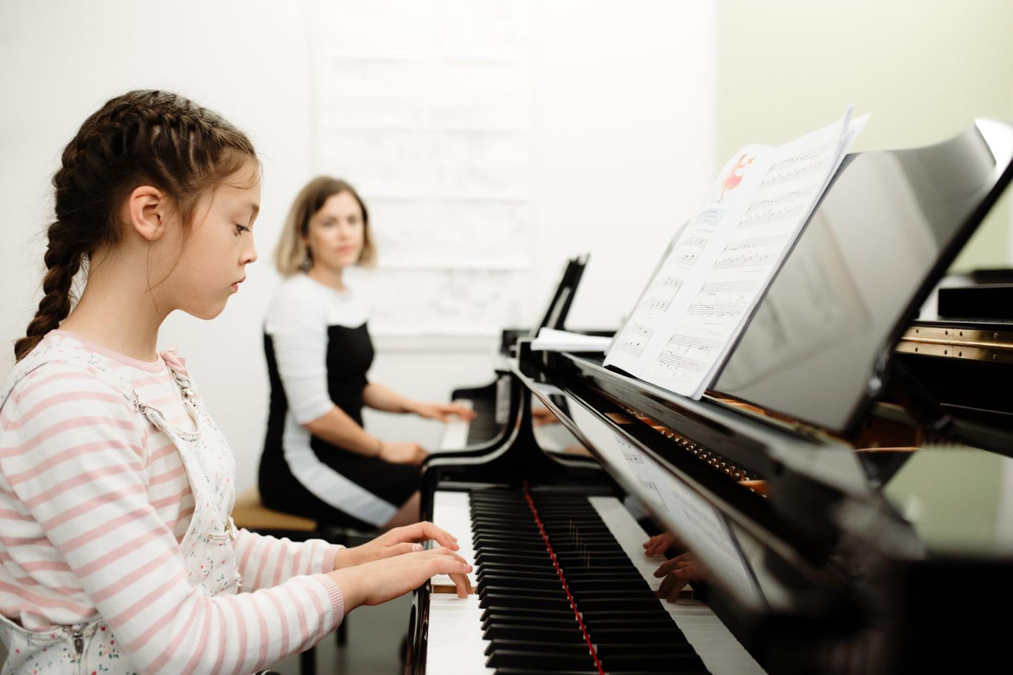 Jong meisje speelt piano, naast haar in de achtergrond staat nog een piano waar een vrouw zit en toekijkt.
