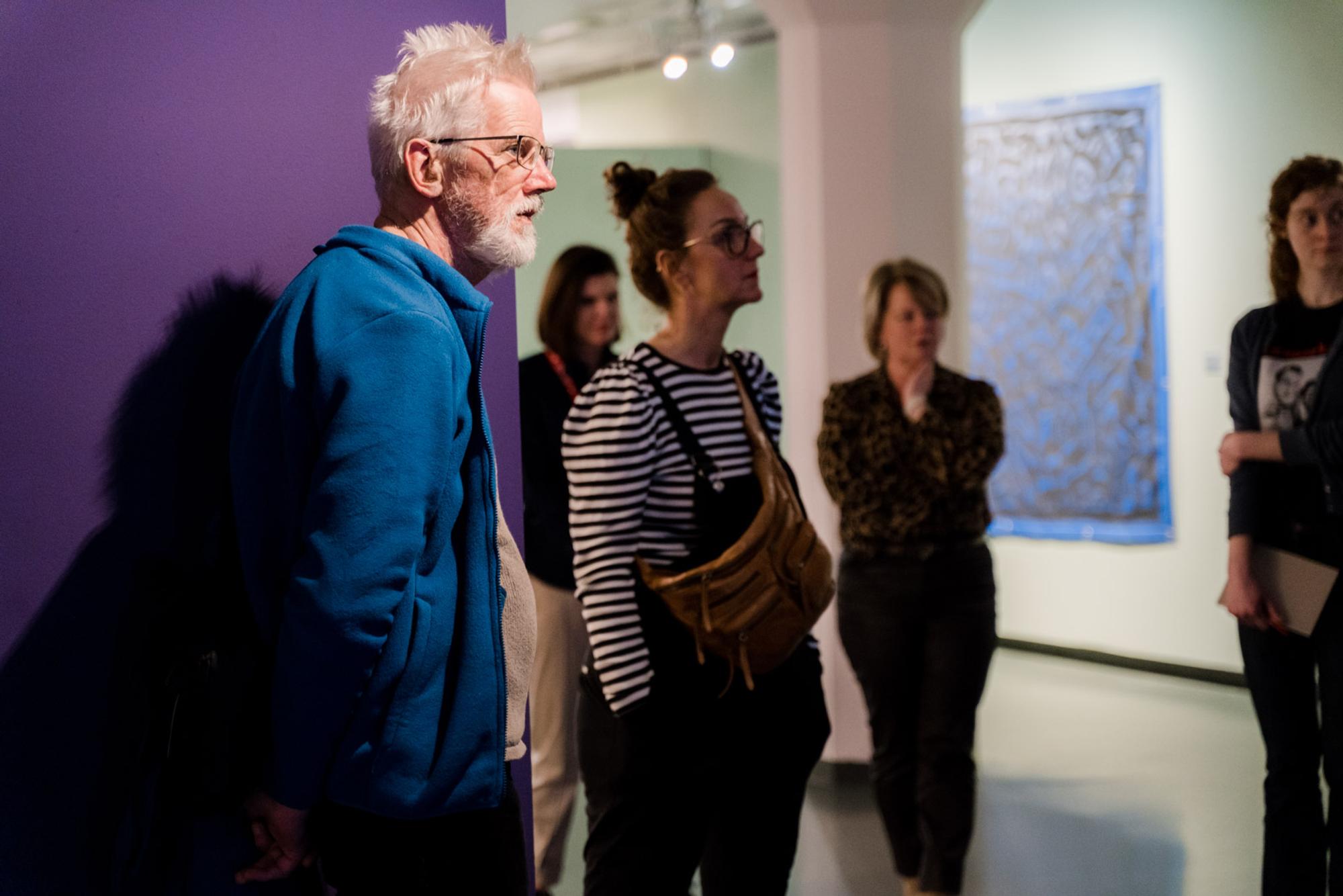Een groep mensen staat aandachtig te luisteren in een tentoonstellingsruimte; op de voorgrond een oudere man met wit haar en blauwe fleecejas, op de achtergrond een kunstwerk aan de muur en paarse wand links in beeld.