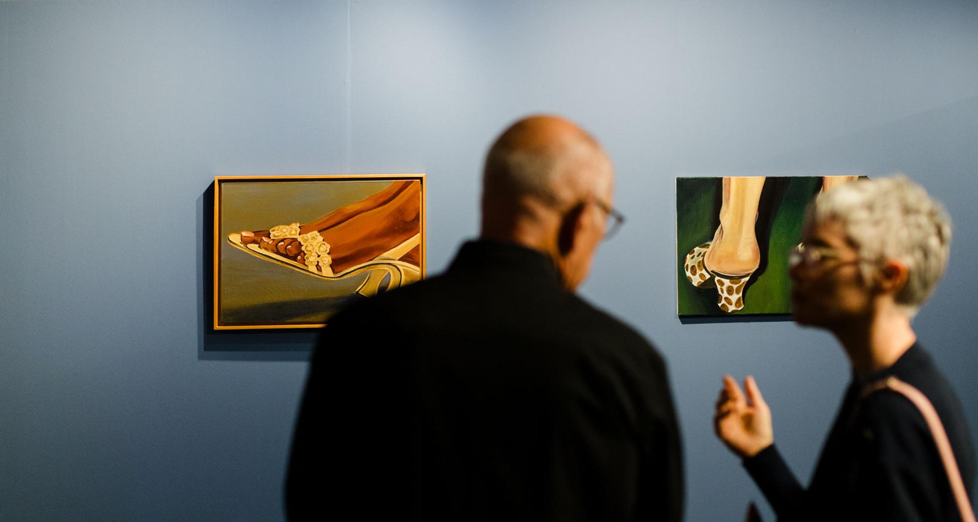 Twee bezoekers bekijken en bespreken schilderijen in een tentoonstellingsruimte, terwijl close-ups van voeten in opvallende schoenen aan een blauwe wand hangen.