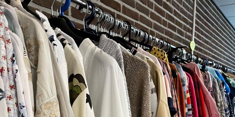 Kleding hangt aan een lange rek met metalen hangers tegen een bakstenen muur. De kledingstukken variëren van lichte en neutrale tinten tot fellere kleuren en verschillende stoffen, waaronder blouses, jurken en truien.