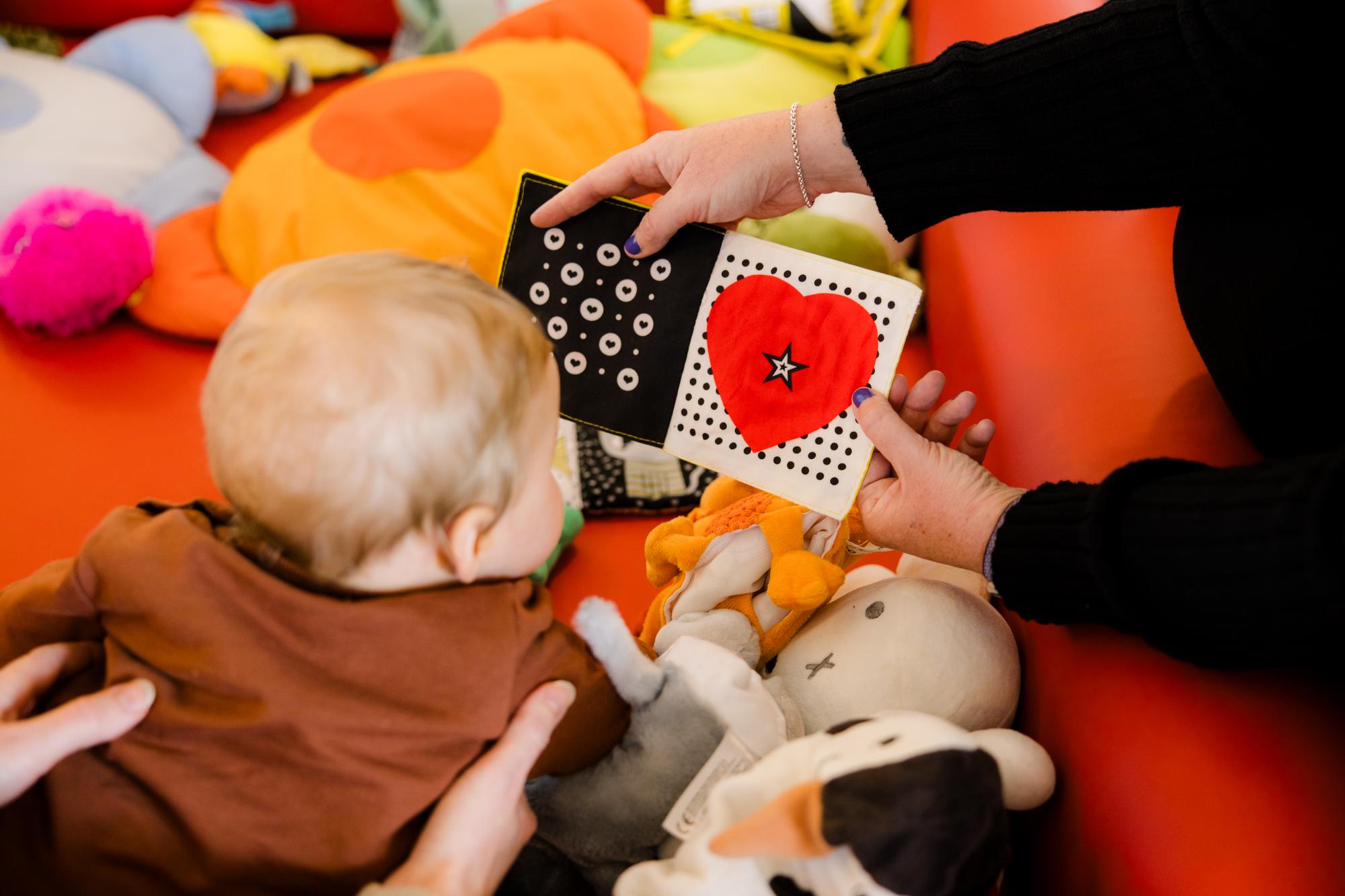 Een baby ligt tussen knuffels op een speelkleed terwijl een volwassene een zwart-wit contrastboekje met een rood hart en stippen voorhoudt.